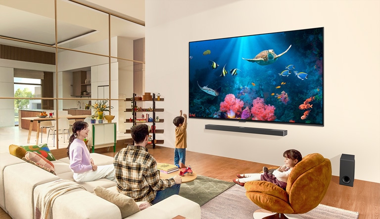 Eine Familie in einem Wohnzimmer mit einem ultragroßen LG-Fernseher an der Wand, auf dem eine Meeresszene mit Korallen und einer Schildkröte abgebildet ist.
