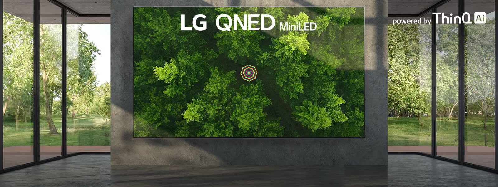 Un televisor LG QNED MiniLED con pantalla ultra grande montado contra una pared con ventanas del piso al techo a cada lado. La pantalla muestra una vista de pájaro de un bosque verde y frondoso.