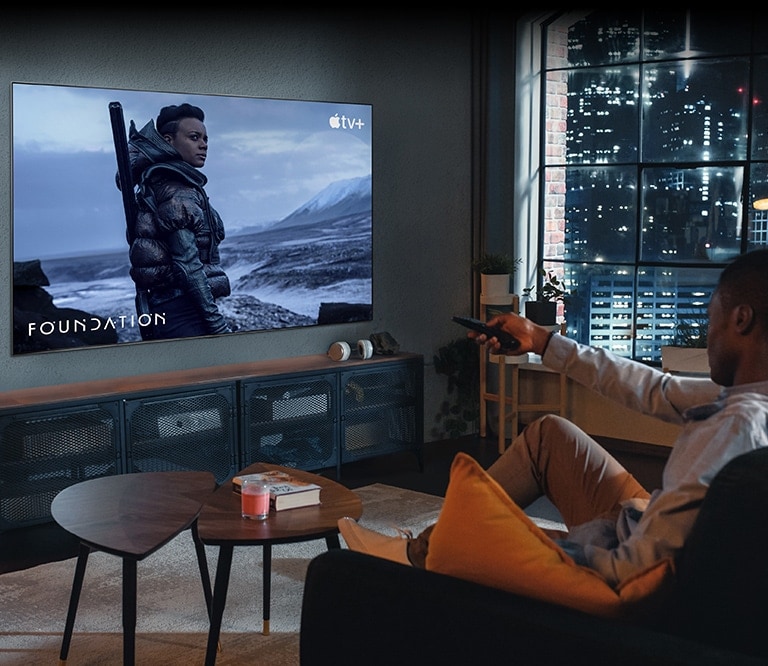 Um homem está sentado no sofá vendo TV. Ele tem um controle remoto na mão, e a TV tem na tela uma cena de “Fundação”, da Apple TV+.