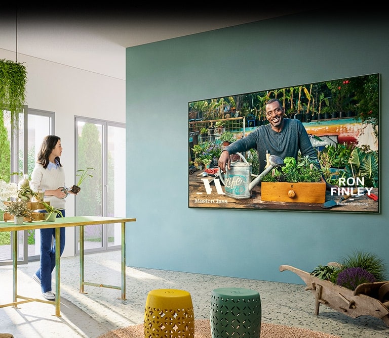 Uma grande TV está na parede, mostrando na tela a aula de jardinagem do “Master Class”. Uma mulher de pé diante da bancada ao lado da TV acompanha a aula manipulando um vaso com planta e uma tesoura de poda.