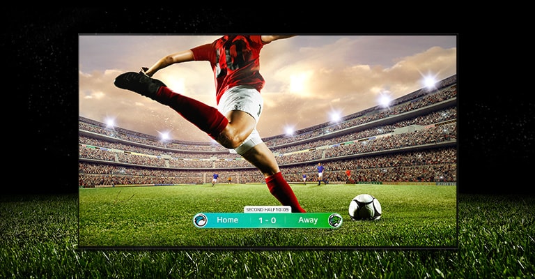 Imagen de la pantalla que muestra un partido de fútbol con un jugador vestido con una franja roja a punto de chutar el balón a través del estadio. El marcador del partido aparece en la parte inferior de la pantalla. El césped verde del campo se extiende más allá de la pantalla hasta el fondo negro.