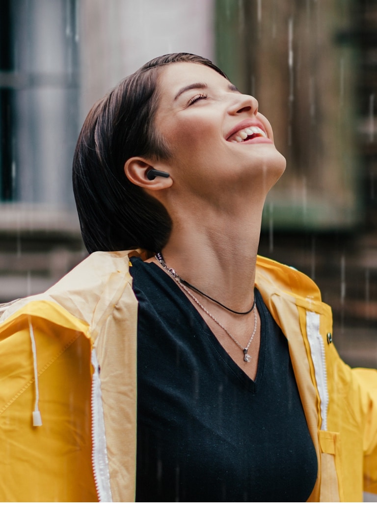 Eine Frau trägt einen farbenfrohen Regenmantel und hört sich etwas über die Ohrhörer an, während Sie im Regen steht.