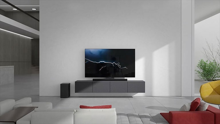 La sound bar è posizionata su un mobile grigio con un TV in salotto. Un subwoofer wireless nero è posizionato sul pavimento sul lato sinistro e la luce del sole entra dal lato destro dell'immagine. Un divano lungo di colore bianco e rosso è posizionato di fronte al TV e alla sound bar.