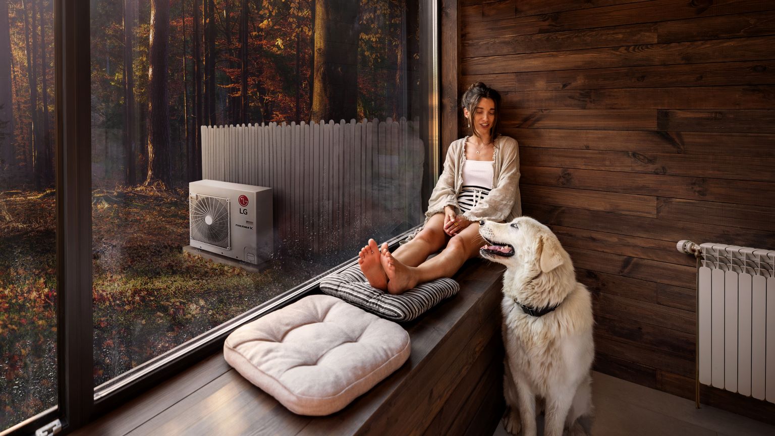 Riscaldamento domestico e soluzioni sostenibili: immagine di una donna con un cane in casa riscaldata dalla pompa di calore aria-acqua LG Thelma V all’esterno.    