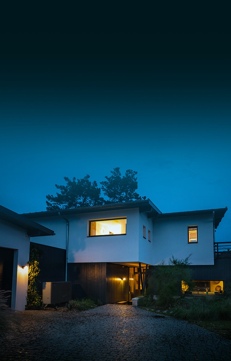 In una fredda serata invernale, l’esterno di una casa con le luci calde accese. La nuova pompa di calore aria-acqua THERMA V LG, di colore nero, è posizionata davanti alla casa. 