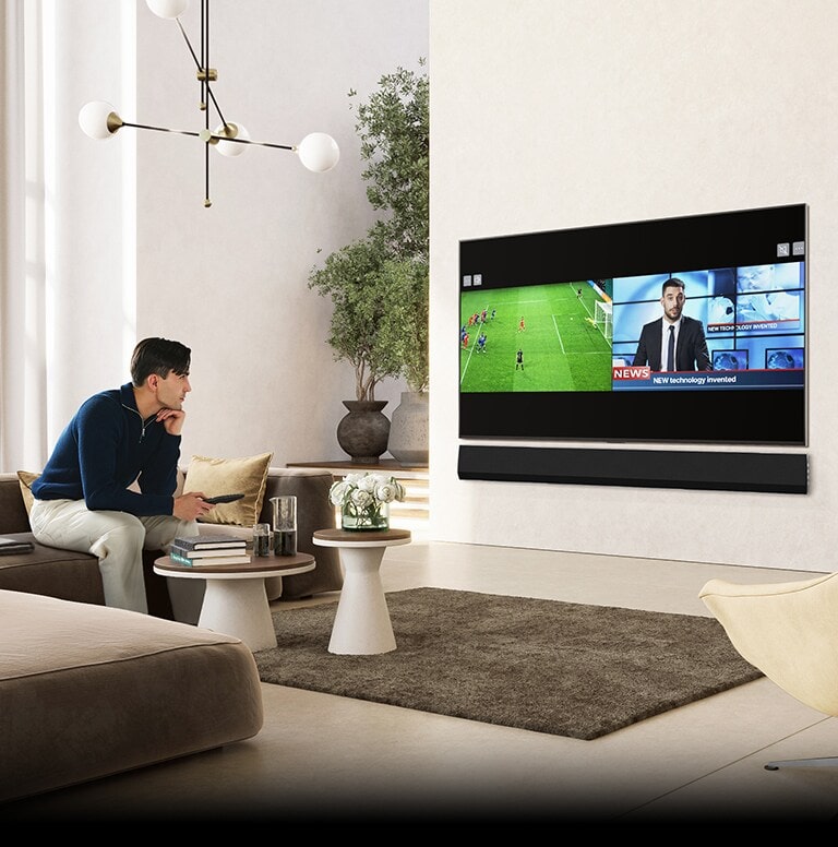 Un uomo è accomodato su un divano in un salotto lussuoso e spazioso e sta guardando il televisore. Sullo schermo del televisore viene visualizzata una schermata divisa tra una partita di calcio e un notiziario.