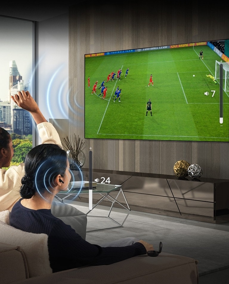 Un gruppo di persone è seduto su un divano a guardare una partita di calcio in TV. La donna sulla destra indossa gli auricolari e li usa con un volume diverso da quello del televisore, ciò indica che li sta usando entrambi contemporaneamente.