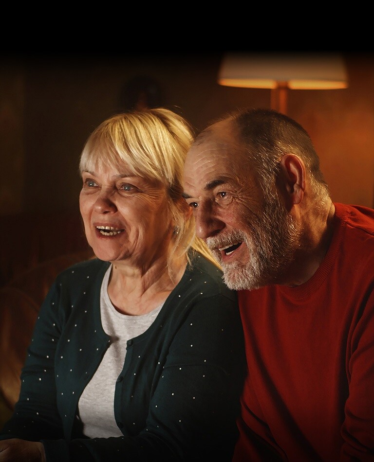 Una coppia di anziani è seduta su un divano e sorride mentre guarda lo schermo.