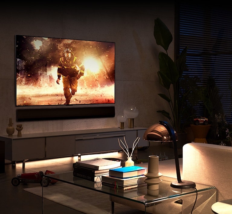 In un salotto spazioso e confortevole, un televisore e una soundbar sono montati a parete. Sullo schermo del televisore si vede un vigile del fuoco che salta fuori da un edificio in fiamme.