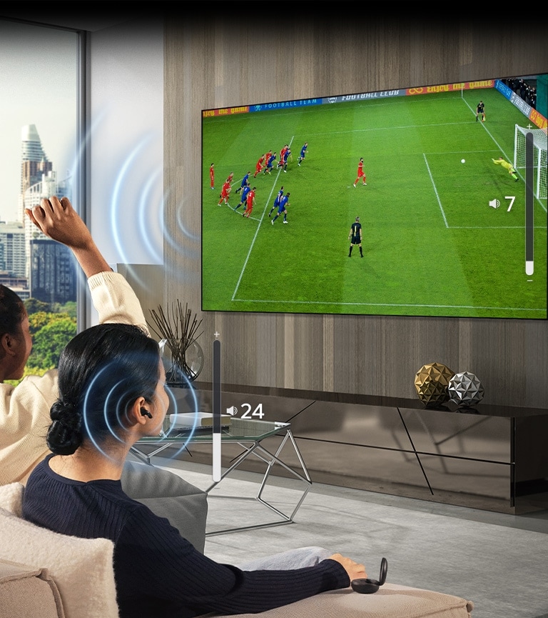 Un gruppo di persone è seduto su un divano a guardare una partita di calcio in TV. La donna completamente a destra indossa gli auricolari e li usa con un volume diverso rispetto a quello del televisore, a indicare che li sta usando entrambi contemporaneamente.