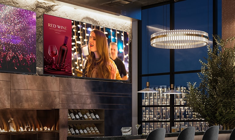 En el lujoso bar de vinos hay dos expositores. Uno muestra una escena de concierto y el otro muestra dos imágenes en una pantalla que muestran un anuncio comercial de vino tinto y una cantante cantando.