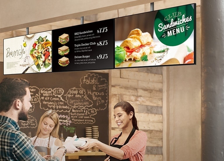 El personal de una tienda de sándwiches está entregando un sándwich a un cliente. La serie SM5J que muestra un tablero de menú está instalada encima de ellos, mostrando menús de sándwich con promociones de brunch.