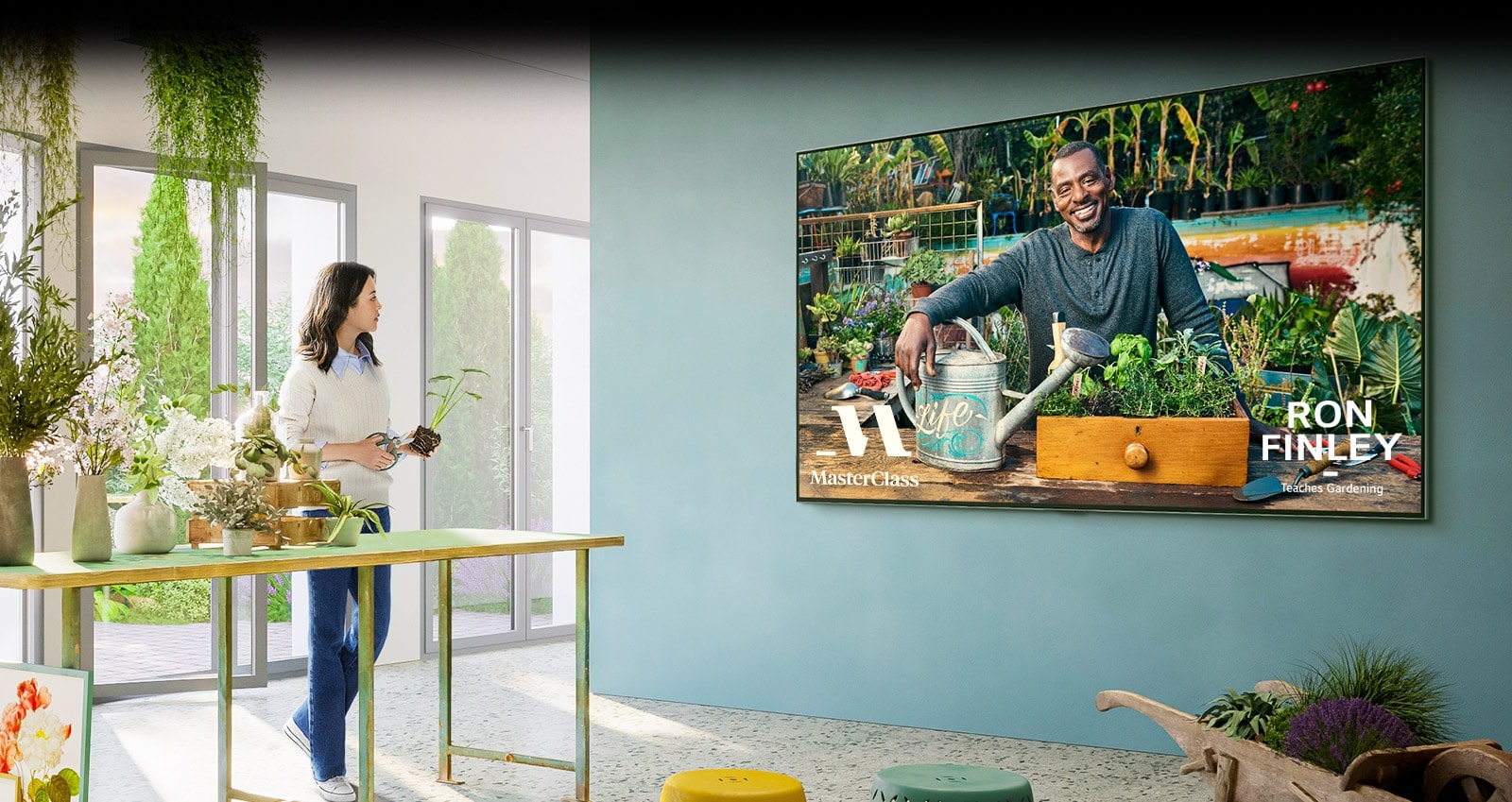  Uma TV grande está na parede e a TV mostra o ecrã da aula de jardinagem de 'Master Class'. Uma mulher está junto a uma mesa ao lado de uma TV e está a assistir a uma aula de jardinagem com vasos de flores e uma tesoura de podar.