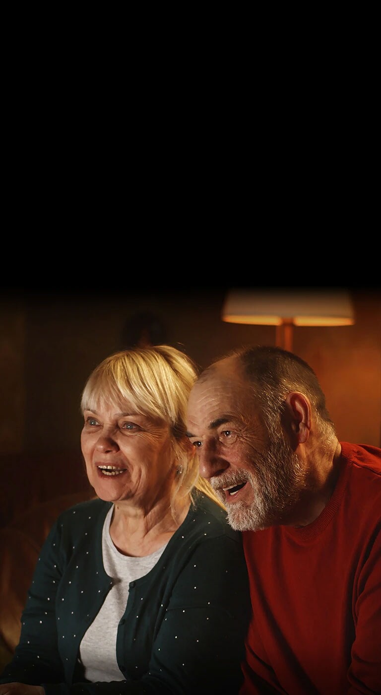 Um casal idoso está sentado num sofá, a sorrir e a olhar para o ecrã.