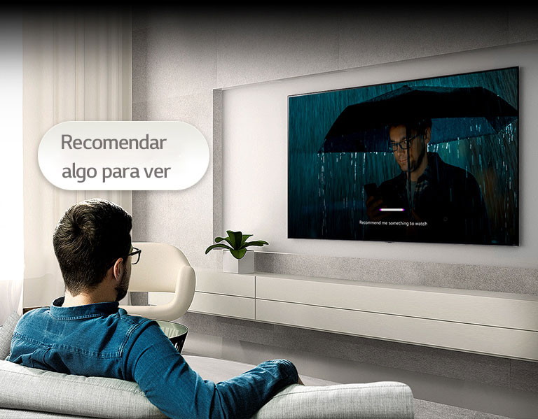  Um homem está sentado num sofá de frente para uma TV no lado oposto. Um balão de fala que diz 'Recomenda algo para ver' está por cima da sua cabeça. No ecrã, está a ser reproduzido o vídeo de um homem a usar um guarda-chuva com uma UI com reconhecimento de voz.
