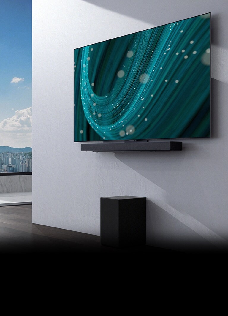  Ao centro de um espaço com uma grande janela está uma parede com uma TV e uma barra de som montada na mesma e um subwoofer por baixo. O ecrã apresenta uma imagem de fundo azul.