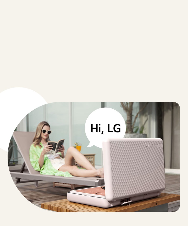 Rear view of the LG StanbyME Go, and it's placed right in front of the patio table. A woman is chilling out on the beach chair, control the screen with her voice. To illustrate this, a speech bubble with Hi, LG text is shown on the right side of her.