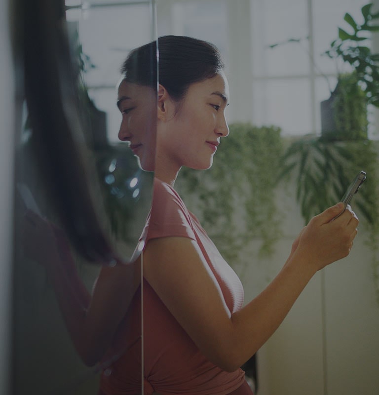 Çamaşır makinesinin yanında duran ve gülümseyerek telefonuna bakan bir kadının görüntüsü.