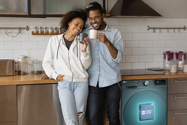 Bardağı tutan çift çamaşır makinesinin yanında ayakta duruyor. AIDD logolu çamaşır makinesin içinden bir AI satırı çıkıyor ve kıyafetleri algılıyor.