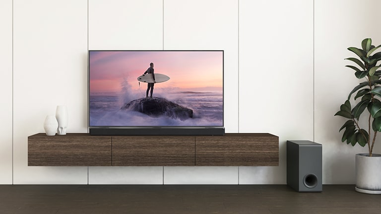 Bir LG TV, LG Soundbar kahverengi bir rafa yerleştirilmiştir ve sub-woofer yerdedir. TV ekranı kayanın üzerinde duran bir sörfçüyü gösterir.