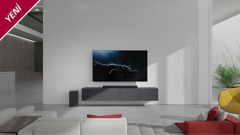 Soundbar, oturma odasında duvara asılı bir TV ile gri dolaba yerleştirilmiştir. Sol taraftaki zemine kablosuz bir subwoofer yerleştirilmiştir ve resmin sağ tarafından güneş ışığı gelir. Beyaz ve kırmızı renkli uzun bir kanepe, TV'ye ve soundbar'a bakacak şekilde yerleştirilmiştir. YENİ işareti sol üst köşede gösterilir.