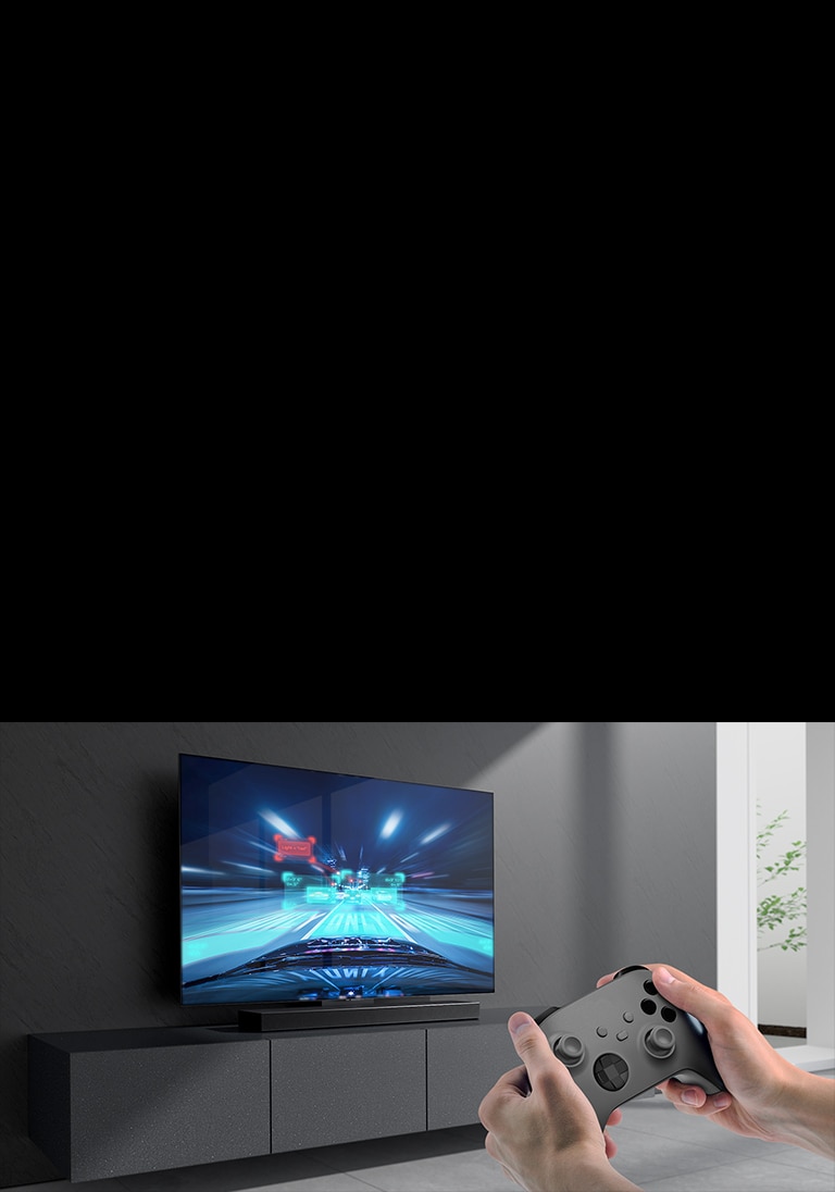Lydplanken er plassert på kabinettet og racingspillscenen vises på TV-en som er koblet til lydplanken. En spillkonsoll er på nedre høyre side av bildet som holdes med to hender.
