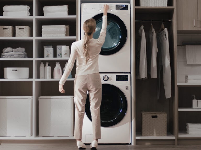 A white washer and dryer are stacked on top of each other and installed in a wall unit with shelves and a closet and a woman has her back facing the front as she reaches up to press a button on the center top control panel. She stands uncomfortably.