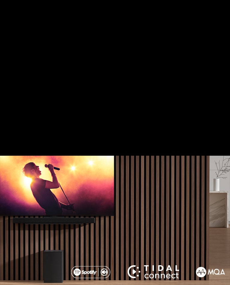Na ścianie wisi telewizor LG OLED serii C, pod nim znajduje się soundbar LG SC9S przymocowany za pomocą specjalnego uchwytu. Pod spodem znajduje się subwoofer. Na ekranie telewizora jest ukazana scena z koncertu.