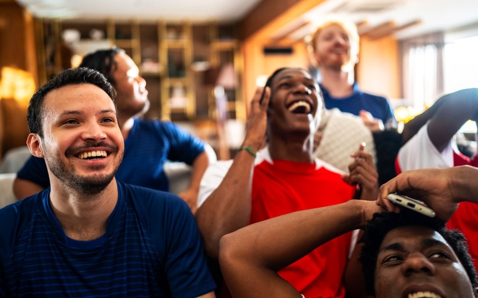 Um grupo de homens assiste alegremente a um jogo de futebol num televisor calibrado, entre risos e sorrisos.