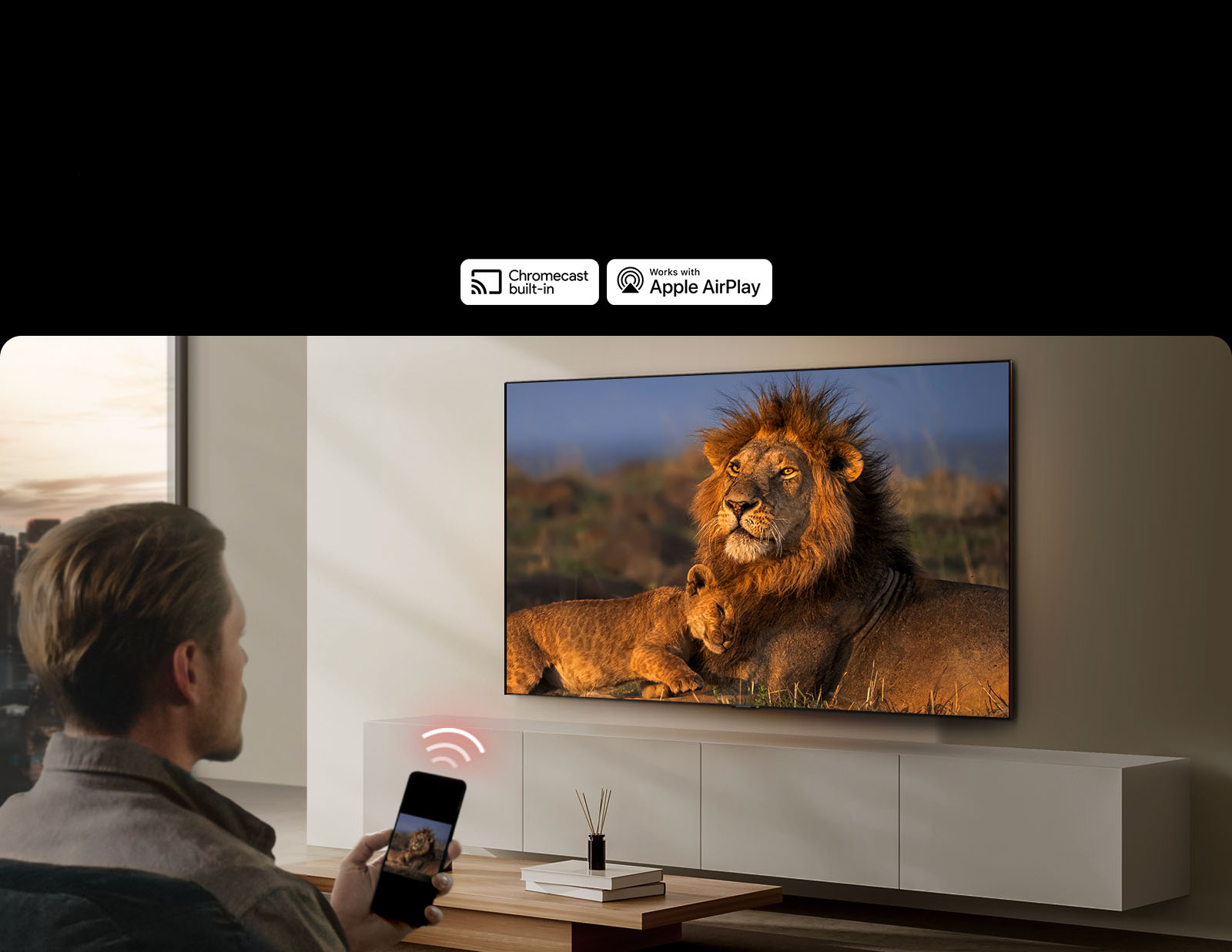 Một chiếc TV LG gắn trên tường trong phòng khách, trưng bày một con sư tử và sư tử con. Một người đàn ông ngồi ở phía trước với chiếc điện thoại thông minh trên tay hiển thị hình ảnh tương tự của những con sư tử. Hình ảnh ba thanh cong màu đỏ neon được hiển thị ngay phía trên điện thoại thông minh hướng về phía TV.
